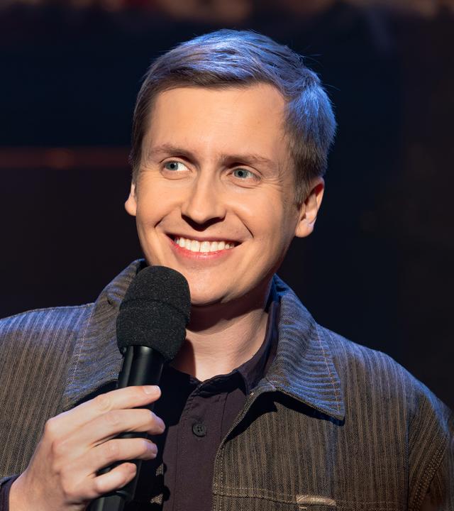 Till Reiners ist auf der rechten Seite des Bildes abgebildet. In seiner rechten Hand hält er ein Mikrofon. Reiners lächelt. Der Hintergrund ist dunkel.