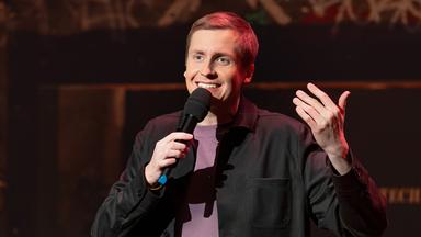 Till Reiners ist in der Mitte des Bildes abgebildet. In seiner rechten Hand hält er ein Mikrofon. Reiners lächelt. Der Hintergrund ist dunkel. Er hebt seine linke Hand. 