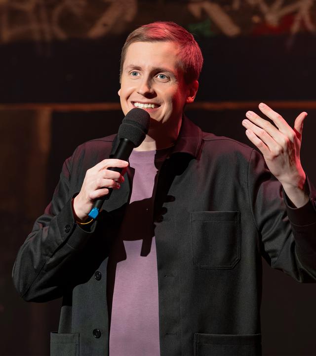 Till Reiners ist in der Mitte des Bildes abgebildet. In seiner rechten Hand hält er ein Mikrofon. Reiners lächelt. Der Hintergrund ist dunkel. Er hebt seine linke Hand. 