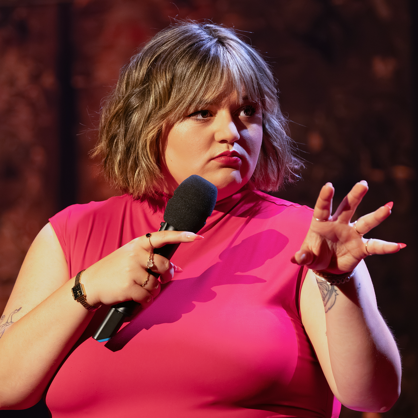 Teresa Reichl steht in der Mitte des Bildes. Sie hält ein Mikrofon in ihrer rechten Hand. Ihre linke Hand hält sie mit gespreizten Fingern in Richtung des rechten Bildrandes. Ihr Oberteil ist rot.