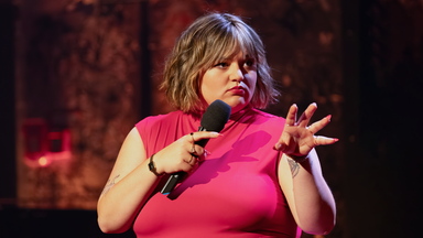 Teresa Reichl steht in der Mitte des Bildes. Sie hält ein Mikrofon in ihrer rechten Hand. Ihre linke Hand hält sie mit gespreizten Fingern in Richtung des rechten Bildrandes. Ihr Oberteil ist rot.