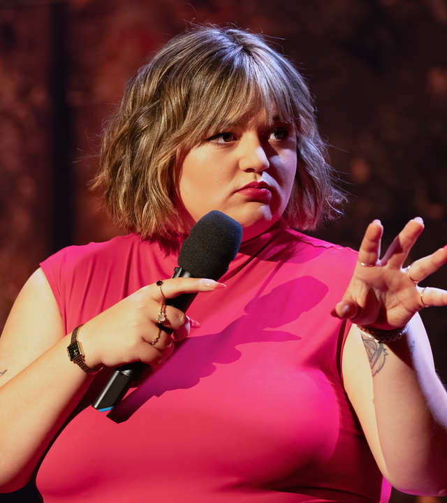 Teresa Reichl steht in der Mitte des Bildes. Sie hält ein Mikrofon in ihrer rechten Hand. Ihre linke Hand hält sie mit gespreizten Fingern in Richtung des rechten Bildrandes. Ihr Oberteil ist rot.