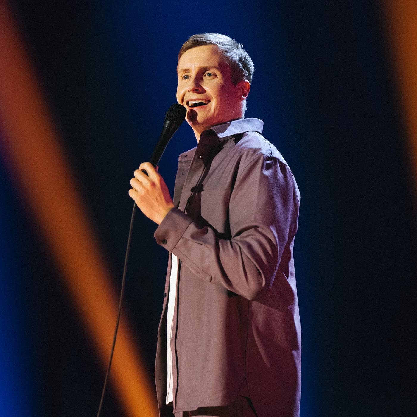 Till Reiners steht auf der rechten Bildseite. In seiner linken Hand hält er ein Mikrofon. Der Hintergrund ist blau und orange beleuchtet.