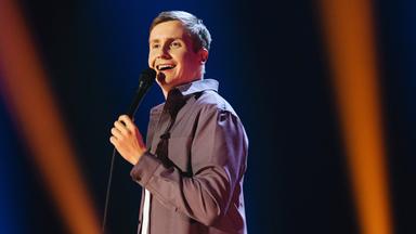 Till Reiners steht auf der rechten Bildseite. In seiner linken Hand hält er ein Mikrofon. Der Hintergrund ist blau und orange beleuchtet.