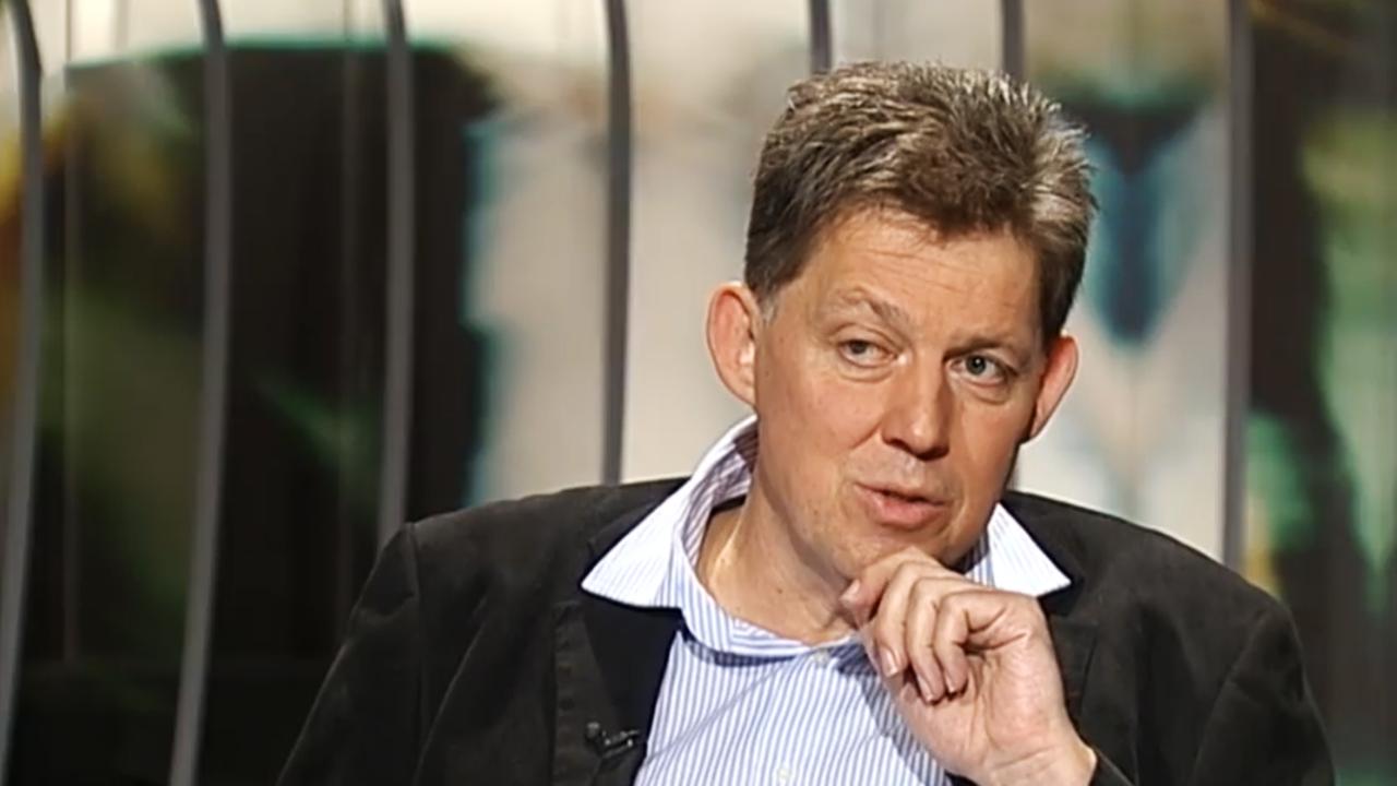 Halbtotale eines Mannes in Hemd und Anzug, der sich beim Reden eine Hand ans Kinn hält.