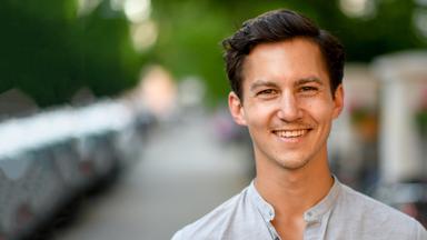 Ein junger Mann vor verschwommenem grünen Hintergrund lacht in die Kamera.