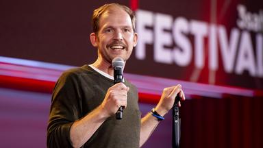 Timo Wopp steht in der Mitte des Bildes. Mit seiner linken Hand hält er einen Mikrofonständer. In seiner rechten Hand hält er ein Mikrofon. Der Hintergrund ist rot beleuchtet.