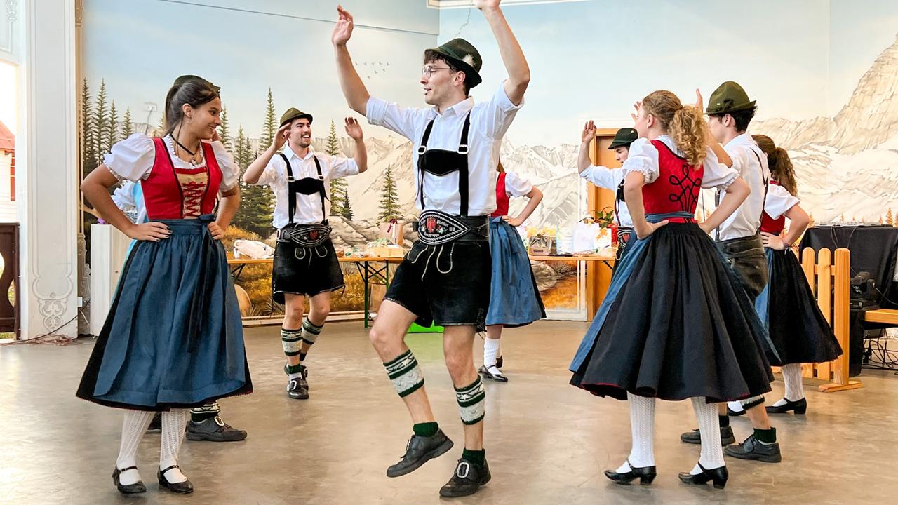 Auf dem Bild ist eine Gruppe von Tänzern zu sehen, die in traditioneller Tracht auftreten. Die Szenerie scheint sich in einem Veranstaltungsraum zu befinden, der mit einer Wandmalerei eines Bergpanoramas dekoriert ist.   Die Tänzerinnen tragen lange, dunkle Röcke und blusenähnliche Oberteile, während die männlichen Tänzer kurze Hosen und Hemden mit suspenders (Hosenhaltern) tragen. Einige Männer tragen traditionelle Hüte.   Die Tänzer bewegen sich aktiv und sind in verschiedenen Posen, die Freude und Energie ausstrahlen. Im Vordergrund sind auch einige Requisiten zu erkennen, wie ein Tisch und technische Geräte, die möglicherweise für die Musik oder die Aufnahme des Auftritts verwendet werden.   Die Atmosphäre wirkt festlich und die Tanzperformance scheint eine kulturelle Darbietung zu sein, die die Traditionen der Tiroler Kultur in ein brasilianisches Umfeld integriert.