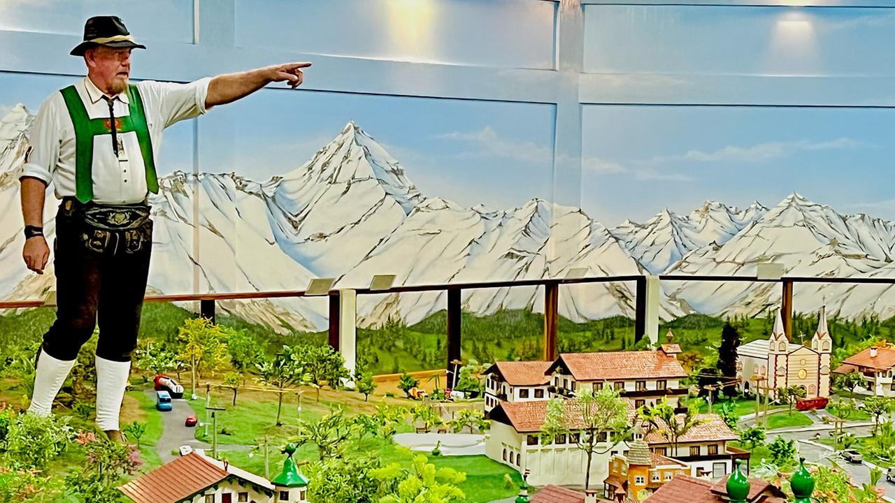 Das Bild zeigt einen Mann in traditioneller alpiner Tracht, bestehend aus einem weißen Hemd mit grünem Schultergurt und dunkler Hose. Er steht auf einer erhöhten Fläche und zeigt mit seinem rechten Arm auf etwas außerhalb des Bildes. Im Hintergrund ist eine große Wandmalerei mit schneebedeckten Bergen zu sehen.  Vor dem Mann befindet sich eine detaillierte Miniaturlandschaft, die Häuser im alpenländischen Stil darstellt, eingerahmt von Bäumen und einem Fahrweg. Die Miniaturgebäude haben rot eingedeckte Dächer und einige von ihnen verfügen über grüne Kuppeln.   Die gesamte Szene vermittelt den Eindruck eines kleinen alpenländischen Dorfes in einer tropischen Umgebung, was den kulturellen Einfluss von Österreich in Brasilien zeigt.