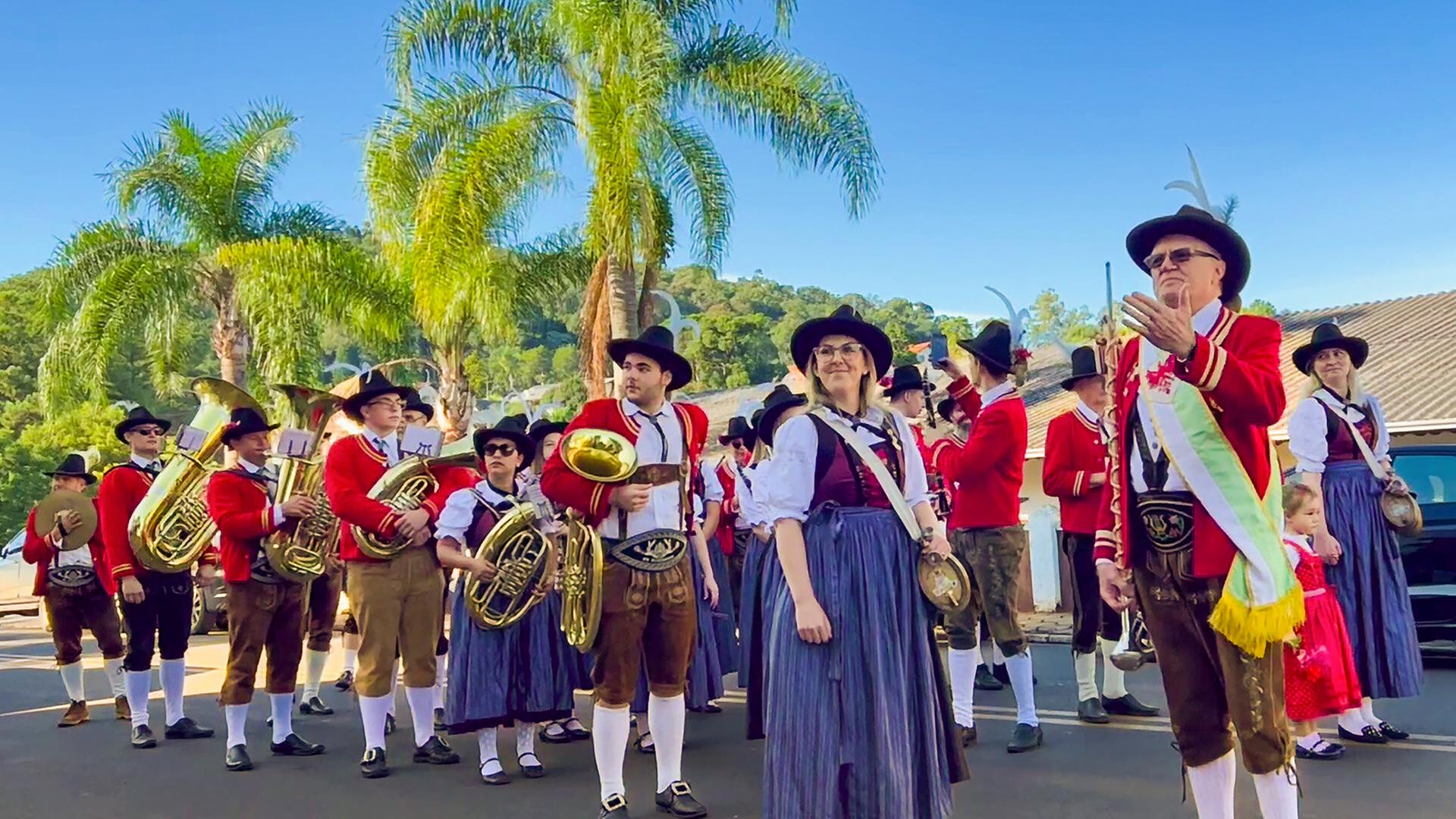 Das Bild zeigt eine Blasmusik-Kapelle in einer festlichen Umgebung. Im Vordergrund stehen mehrere Musiker in traditionellen Trachten mit roten Jacken und breiten Hüten, die mit Federn geschmückt sind. Einige von ihnen halten Instrumente wie Tuba und Trompete. Die meisten tragen Hosen in Erdtönen, kombiniert mit weißen Socken.   Eine auffällig gekleidete Person mit Brille steht in der Mitte des Bildes und hat eine auffallende Schärpe umgelegt. Hinter den Musikern sieht man eine Reihe von Palmen, die auf das warme Klima hindeuten. Der Himmel ist strahlend blau, und im Hintergrund sind einige Gebäude, die möglicherweise zur festlichen Veranstaltung oder zur Gemeinde gehören. Die gesamte Szene vermittelt ein Gefühl von Gemeinschaft und kulturellem Erbe.