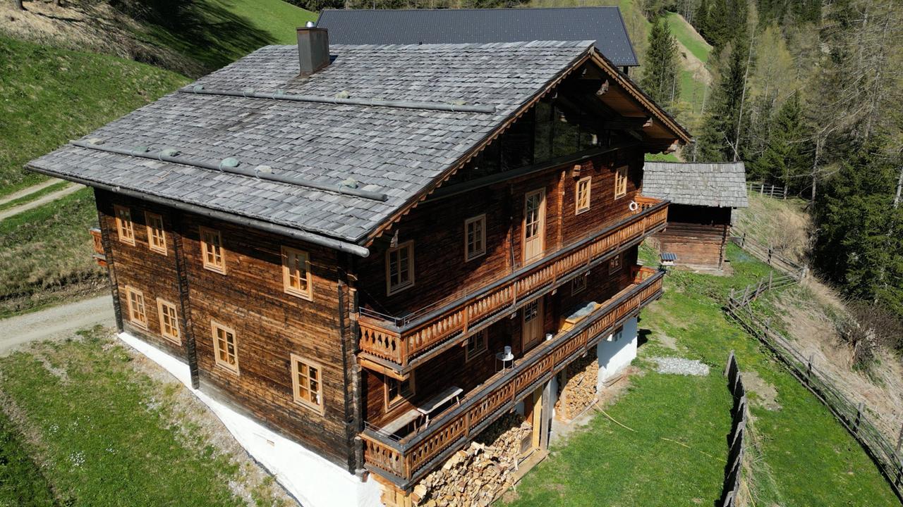 Das Bild zeigt ein traditionelles, mehrstöckiges Tiroler Bauernhaus mit Holzverkleidung und einem steilen, schindelgedeckten Dach. Das Gebäude hat zahlreiche Fenster mit Holzrahmen, die in Reihen angeordnet sind. Im Erdgeschoss ist ein Bereich mit gestapelten Holzscheiten sichtbar, was auf die Nutzung des Gebäudes für landwirtschaftliche Zwecke hinweist.  Rund um das Bauernhaus erstreckt sich eine grüne Wiese, die sanft in eine Hügellandschaft übergeht. Im Hintergrund sind einige Bäume zu erkennen, die vor einer bewaldeten Fläche stehen. Ein Wanderweg führt in der Nähe des Hauses vorbei. Die Umgebung wirkt idyllisch und zeigt die ländliche Kultur und Architektur Tirols.  Der Himmel ist klar und es scheint sonnig zu sein, was die Farben lebendig erscheinen lässt. Das gesamte Bild vermittelt einen Eindruck von traditioneller ländlicher Idylle und der Verbindung zur Natur.