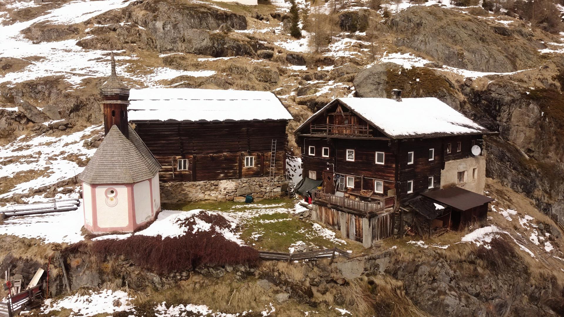 Das Bild zeigt eine ländliche Szenerie in Tirol. Im Vordergrund sind zwei traditionelle Bauernhäuser zu sehen. Das linke Gebäude hat eine helle Fassade mit einem runden, spitzen Dach und einer kleinen, ornamente Stube. An der Wand befindet sich eine dekorative Malerei. Das rechte Gebäude ist aus dunklem Holz konstruiert, hat mehrere Fenster und einen Balkon. Die Dächer beider Häuser sind teilweise mit Schnee bedeckt.  Im Hintergrund ist eine hügelige, felsige Landschaft sichtbar, die ebenfalls mit Schnee bedeckt ist. Der Boden ist größtenteils braun und trocken, und einige Grasflächen sind sichtbar. Eine Leiter lehnt an dem linken Haus, und bis auf einige grasbewachsenen Flächen sind keine weiteren Pflanzen vorhanden. Es ist ein klarer, kalter Tag, was durch die Schneebedeckung und die winterliche Umgebung suggeriert wird.