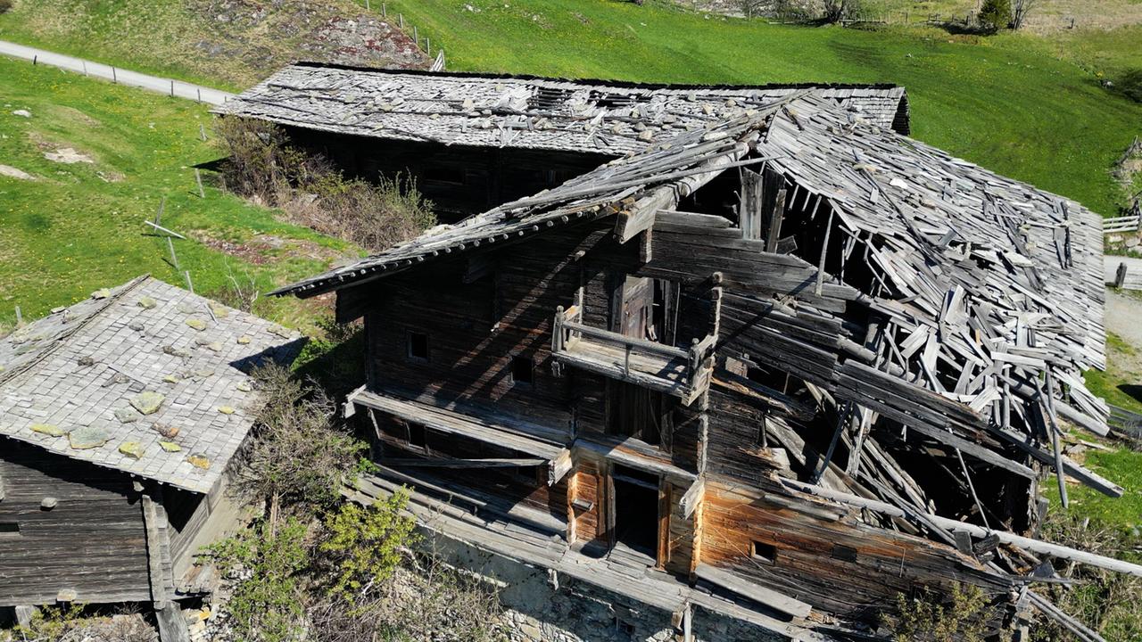 Das Bild zeigt den Bichlerhof in Osttirol, ein stark beschädigtes altes Bauernhaus. Das Gebäude ist aus dunklem Holz und hat ein stark abgenutztes Dach, das teilweise eingestürzt ist. Viele Holzbretter sind schief und liegen lose, während andere Teile des Dachs von Moos und Flechten bedeckt sind. Die Fassaden sind ebenfalls stark verwittert. Um das Haus herum befindet sich eine grüne Landschaft mit niedrigem Gras und vereinzelten wild wachsenden Pflanzen. Im Hintergrund sind sanfte Hügel zu sehen, die die ländliche Umgebung prägen. Der Zustand des Gebäudes reflektiert den Verfall traditioneller Tiroler Bauernhäuser.