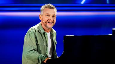 Kabarettist sitzt am Flügel in der Mitte des Bildes. Er trägt ein offenes grünes Hemd. Darunter trägt er ein weißes Shirt. Der Hintergrund ist blau beleuchtet.