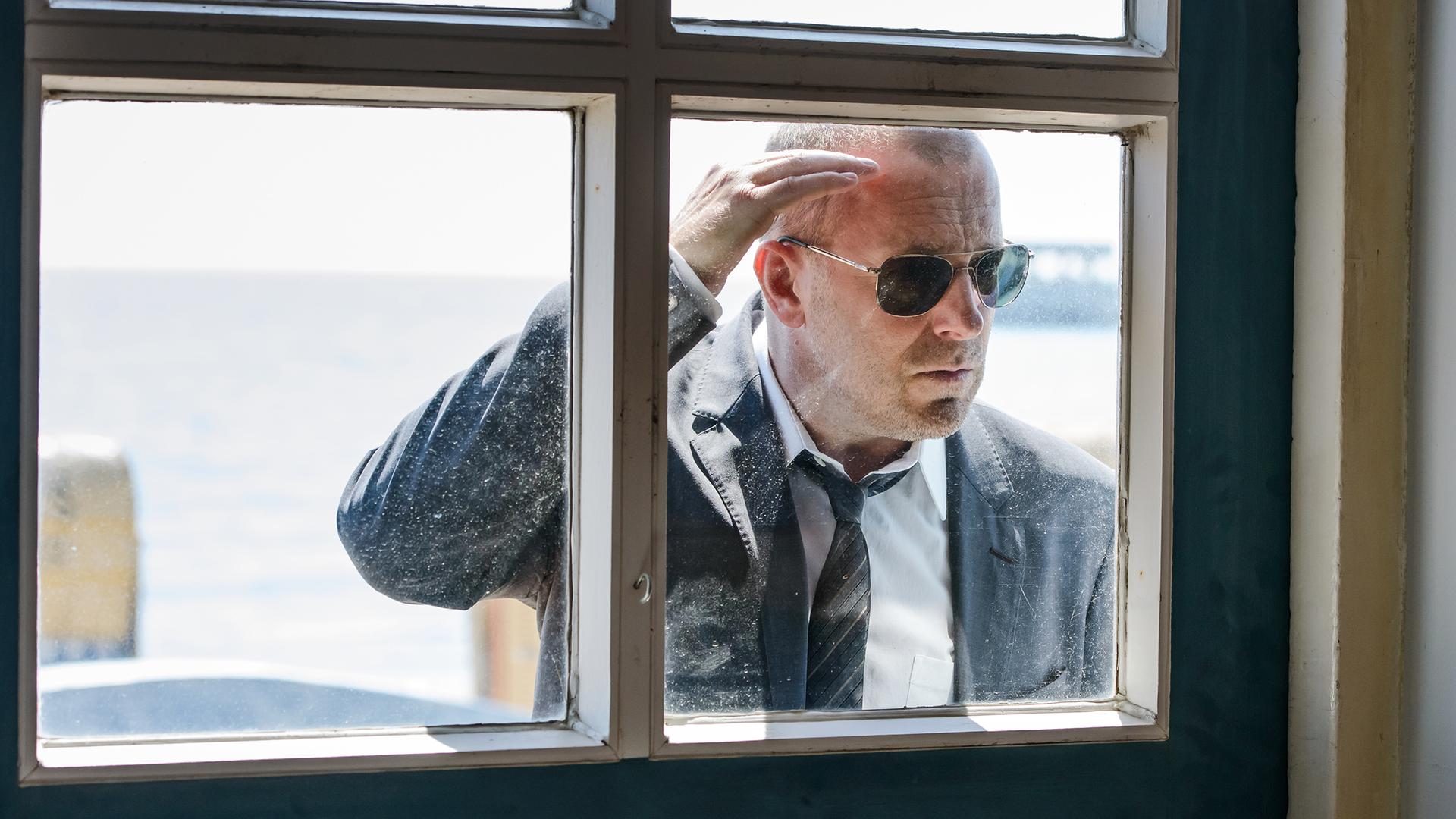 Simon Kessler (Heino Ferch) schaut durch ein Fenster ins Innere eines Wohnhauses. Er trägt einen Anzug mit Krawatte und eine dunkle Sonnenbrille.
