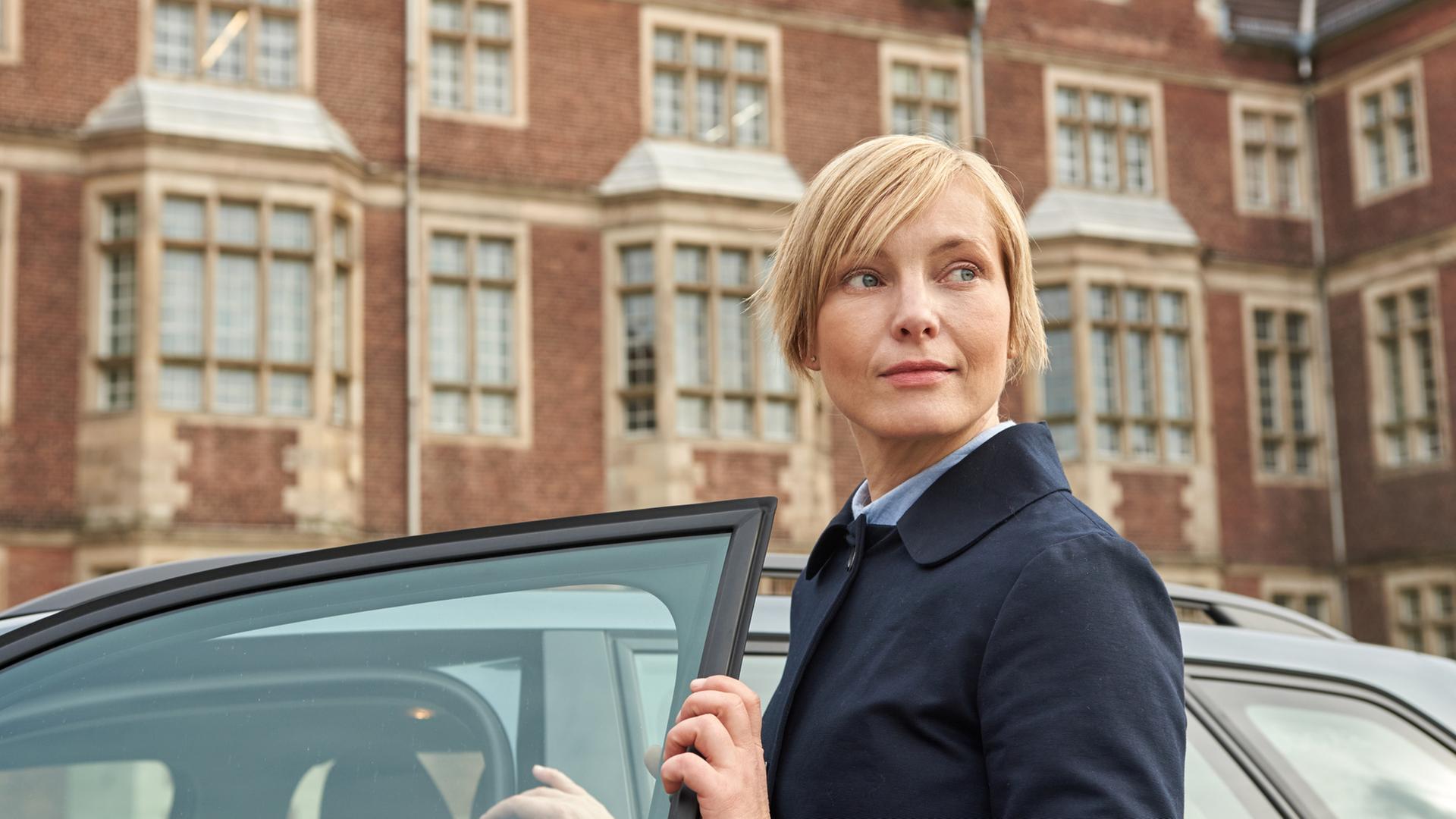"Tod im Internat": Lehrerin Karla Parker (Mitte rechts) ist gerade aus einem Auto ausgestiegen und schaut nun, mit der Autotür in der Hand, in die Ferne. Im Hintergrund ist die Hausfassade des Elite-Internats Erlengrund zu sehen. Tatsächlich ist sie die verdeckte Ermittlerin Isabell Moosbach (Nadja Uhl).