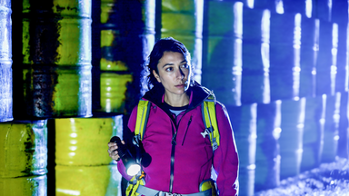 Eine Frau in einer pinkfarbenen Jacke mit Rucksack steht mit einer Taschenlampe in der Hand vor einer Wand aus metallenen Fässern. Sie schaut konzentriert in die Ferne. Im Hintergrund blaues Licht.