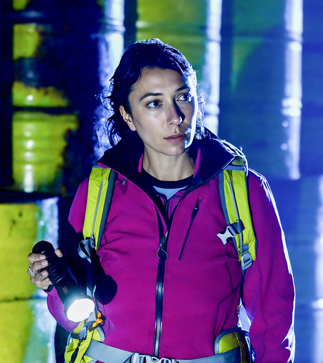 Eine Frau in einer pinkfarbenen Jacke mit Rucksack steht mit einer Taschenlampe in der Hand vor einer Wand aus metallenen Fässern. Sie schaut konzentriert in die Ferne. Im Hintergrund blaues Licht.
