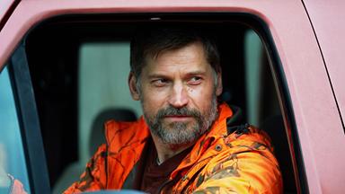 Ein bärtiger Mann mit kurzer Frisur sitzt am Steuer eines Autos und trägt eine orange Tarnjacke. Er blickt ernst aus dem Fenster.