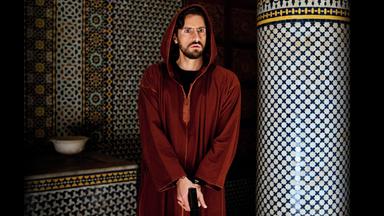 Kommissar Falcón (Juan Diego Botto) steht mit der Pistole in der Hand neben einer Säule und beobachtet entgeistert etwas.