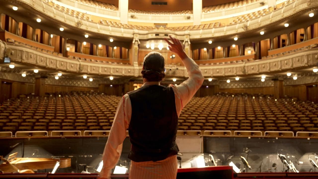 Ein Mann winkt von einer Bühne in einen leeren Opernsaal.