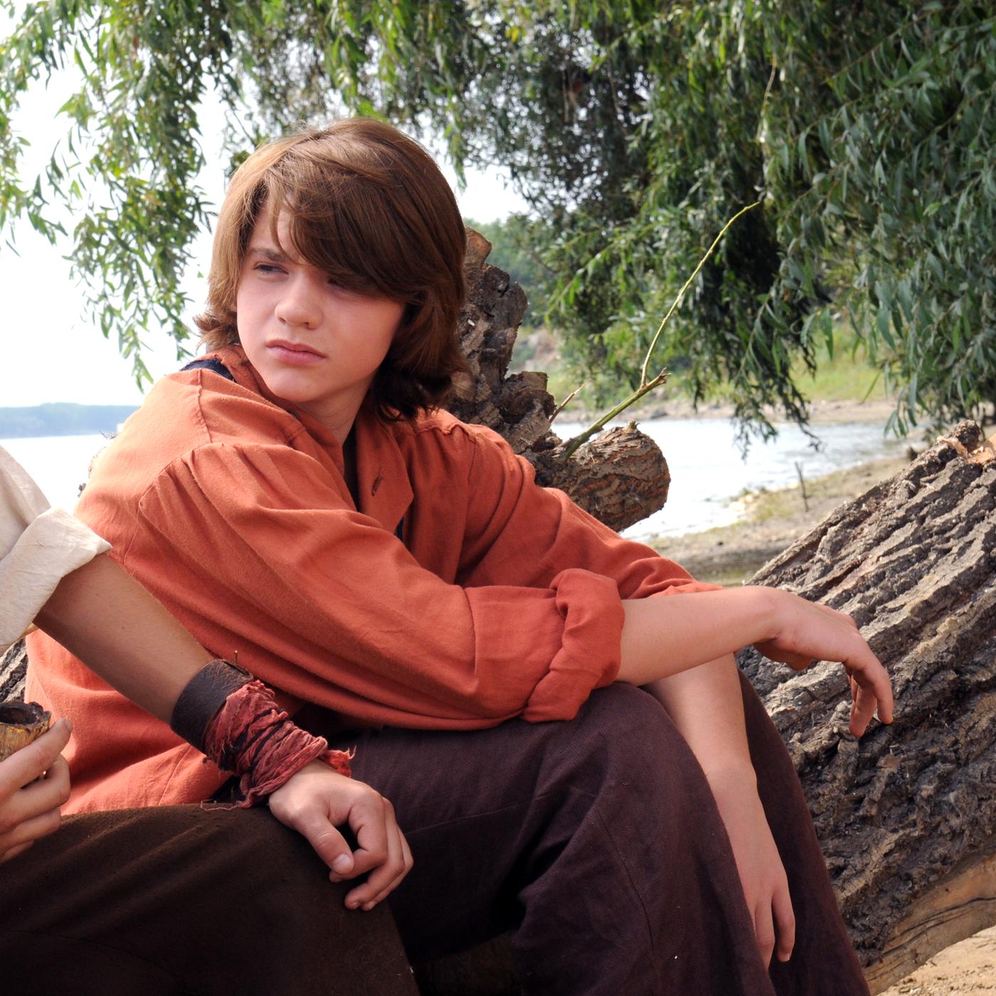 Tom Sawyer (Joel Courtney) und Huckleberry Finn (Jake T. Austin)  sitzen am Ufer des Mississippi auf einem Baumstamm, Tom schaut Huck an, dieser ist mit seiner Pfeife beschäftigt, in der er ständig Maiskolben schmaucht.