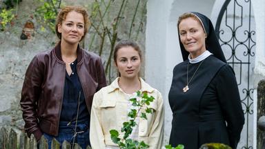 "Tonio & Julia - Dem Himmel so nah": An einem Gartenzaun mit einer Hauswand und einem schmiedeeisernen Tor im Hintergrund stehen Julia Schindel (Oona Devi Liebich), Luise Laibinger (Alina Abgarjan) und Sr. Emanuela (Katharina Meinecke). Alle drei blicken nach vorne.