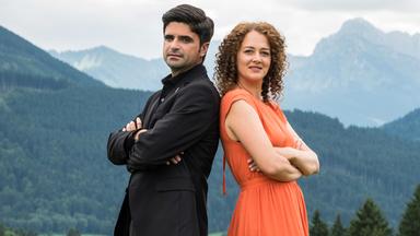 "Tonio & Julia - Kneifen gilt nicht": Mit dem Rücken aneinandergelehnt und nach vorne in die Kamera blickend, stehen Pfarrer Tonio Niederegger (Maximilian Grill) und Familientherapeutin Julia Schindel (Oona Devi Liebich) auf einer Wiese. Im Bildhintergrund Bäume und Berge.