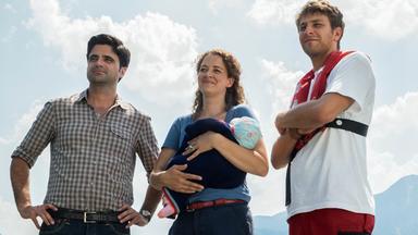 "Tonio & Julia - Zwei sind noch kein Paar": Julia Schindel (Oona Devi Liebich) hält ein Baby im Arm und blickt sehr glücklich. Links und rechts neben ihr stehen Tonio Niederegger (Maximilian Grill) und Guido Bender (Frederik Götz), der eine Rettungsweste trägt. Im Hintergrund blauer Himmel und Berge.