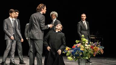 Viele Männer in dunklen Anzügen stehen auf der Bühne. Einer von ihnen hält einem knienden Kirchenküster eine Pistole an den Kopf.