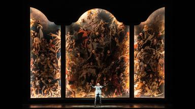 Die Bühne wird von einem dreiteiligen Gemälde, einem Triptychon, eingenommen. Aus dem Bild schauen die Köpfe der Sängerinnen und Sänger vom Chor der Niederländischen Oper in Amsterdam heraus. Am unteren Bildrand steht der Tenor Gevorg Hakobyan in der Rolle des Scarpia.