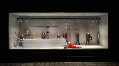 Scarpia und seine Helfer sind in Scarpias Küche. Tosca kniet in einem eleganten roten Kleid auf dem Boden und schaut auf ihren Geliebten Mario Cavaradossi. 