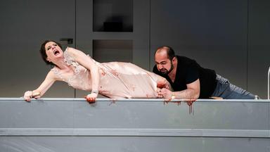 Tosca (Malin Byström) und Scarpia (Gevorg Hakobyan) liegen seitlich auf der Kücheninsel in Scarpias Küche. Tosca ist mit Blut beschmiert.