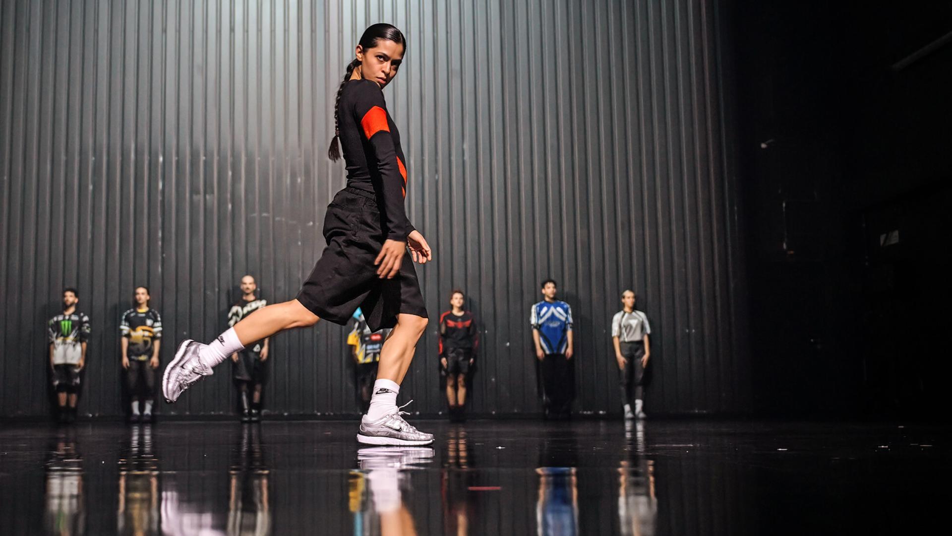 Im Vordergrund eine junge, dunkelhaarige Frau mit einem langen, geflochtenen Zopf, sie steht seitlich und hat das rechte Bein nach hinten angehoben. Sie blickt direkt ins Publikum. Im Hintergrund stehen aufgereiht an der Rückwand der Bühne acht weitere Personen. Alle tragen Motocross Shirts. 