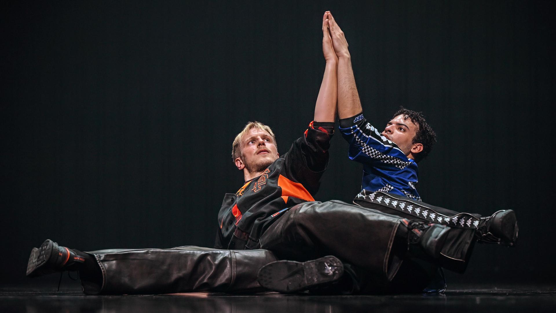 Zwei Tänzer in Motocross Shirts liegen mit gespreizten Beinen seitlich hintereinander auf dem Boden, die Beine ineinander verschränkt, die Oberkörper aufgerichtet, jeweils ihren linken Arm nach oben ausgestreckt aneinandergelegt