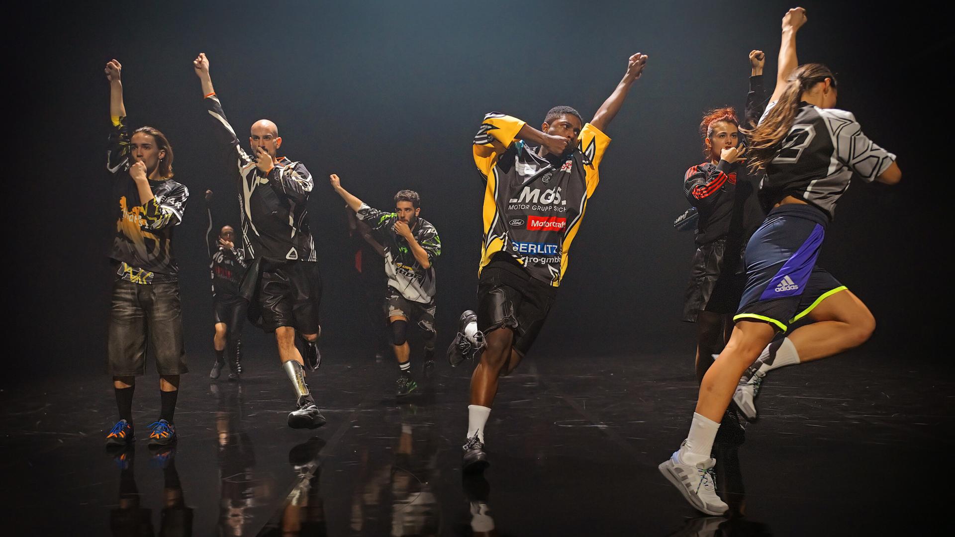 Eine Gruppe von acht Tänzerinnen und Tänzern in Motocross Shirts läuft Richtung Bühnenkante. Sie haben alle eine Faust in die Luftgestreckt und die andere zum Mund geführt. 