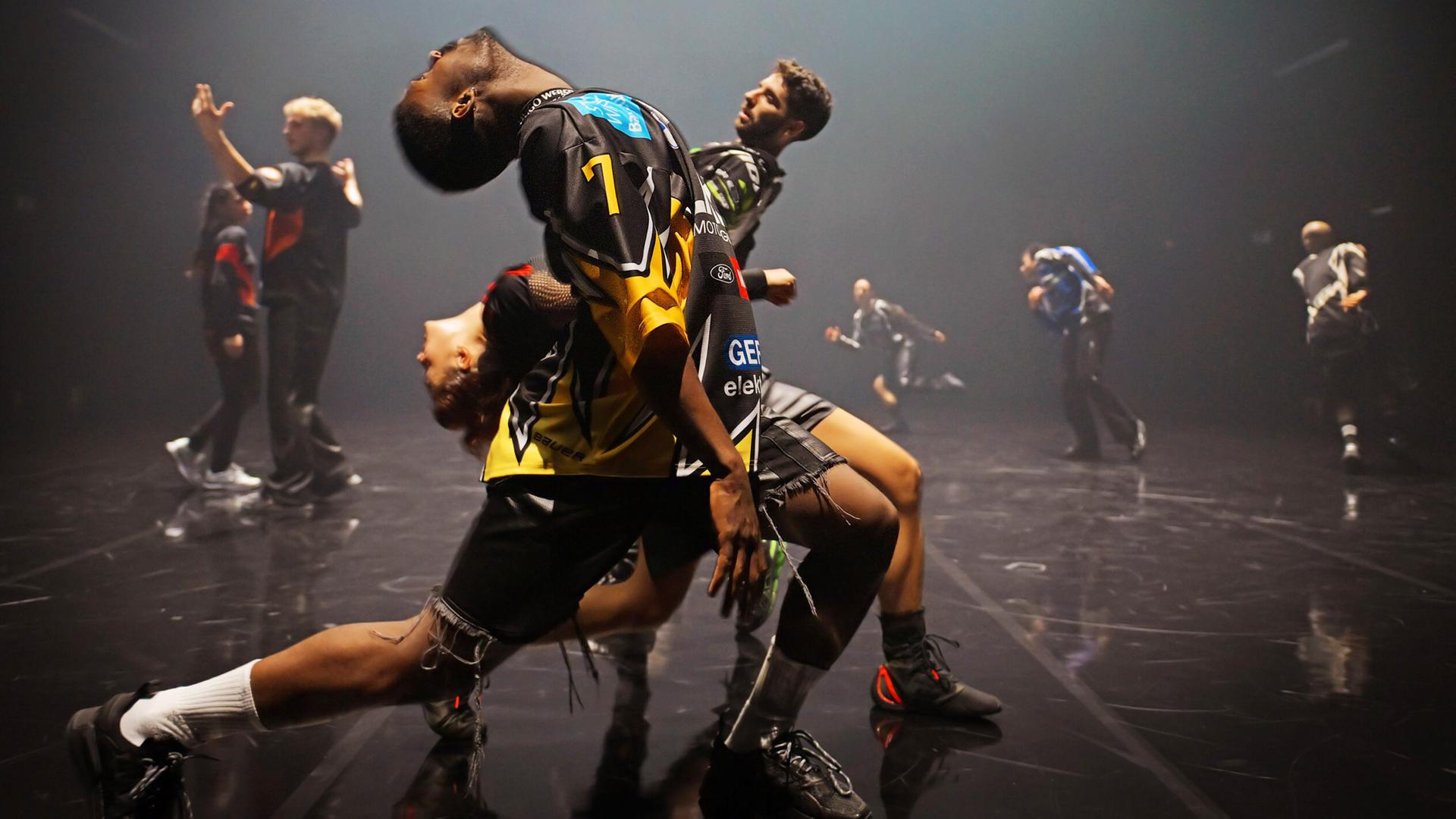 Zwei Tänzer und eine Tänzerin stehen seitlich zum Publikum hintereinander in einem weiten Ausfallschritt und beugen ihre Oberkörper nach hinten. Der vordere Tänzer und die Tänzerin nach links, der hinterste Tänzer nach rechts. Im Hintergrund sind fünf weitere Tänzerinnen und Tänzer zu erkennen.  