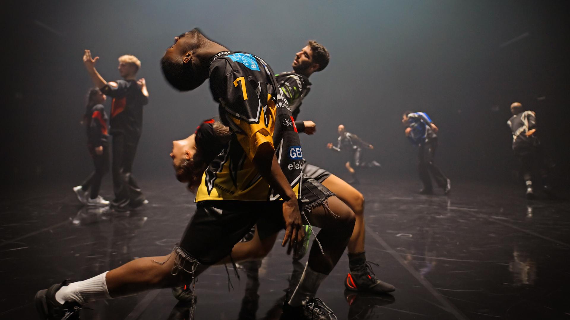 Zwei Tänzer und eine Tänzerin stehen seitlich zum Publikum hintereinander in einem weiten Ausfallschritt und beugen ihre Oberkörper nach hinten. Der vordere Tänzer und die Tänzerin nach links, der hinterste Tänzer nach rechts. Im Hintergrund sind fünf weitere Tänzerinnen und Tänzer zu erkennen.  