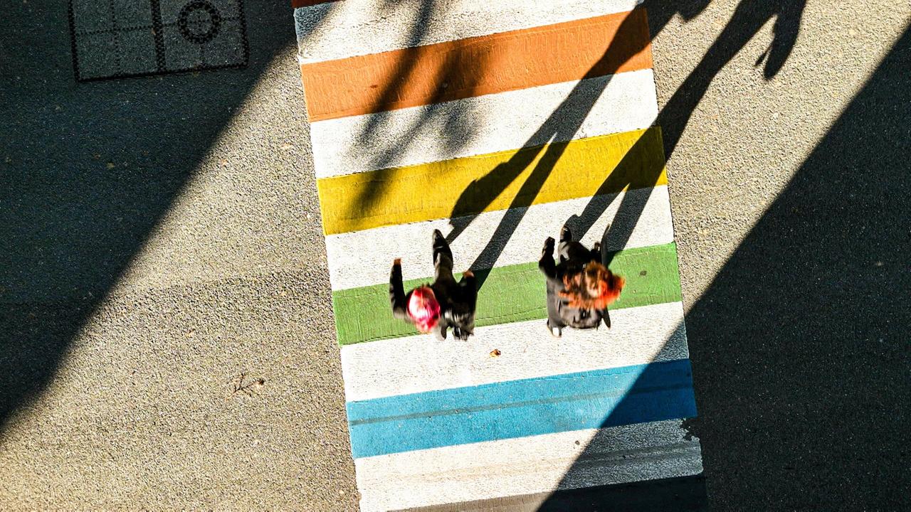 Zwei Personen überqueren einen Zebrastreifen, der in den Farben der Pride-Flagge bemalt ist. Die Aufnahme zeigt die Szene aus der Vogelperspektive, die langen Schatten der Personen fallen auf die Straße.