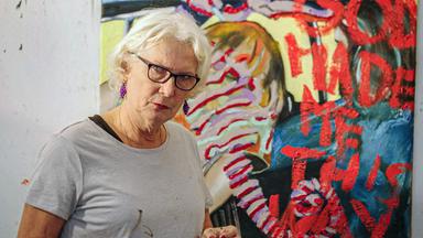 Cornelia Kunert steht in ihrem Atelier vor einem farbenfrohen Gemälde mit abstrakten Elementen und roten Schriftzügen.