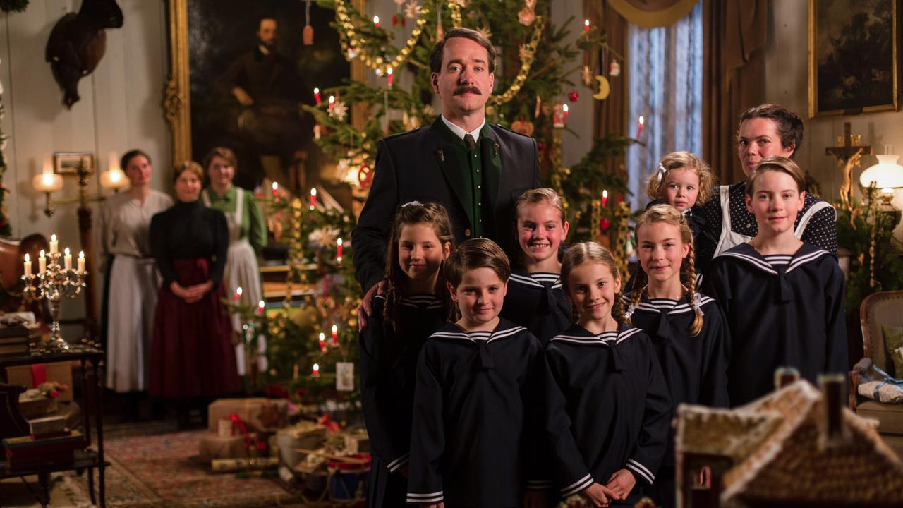 Das Bild zeigt eine Gruppe von Personen in einem festlich dekorierten Raum, um einen Weihnachtsbaum versammelt. Im Vordergrund stehen sechs Kinder in Marineanzügen und ein Mann. Im Hintergrund sind weitere Personen und Einrichtungselemente zu sehen.