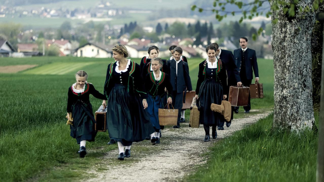 Gruppe von Menschen, die auf einem Feldweg laufen. Sie tragen traditionelle Kleidung und haben Gepäck dabei. Im Hintergrund sind grüne Wiesen und vereinzelte Häuser sichtbar.