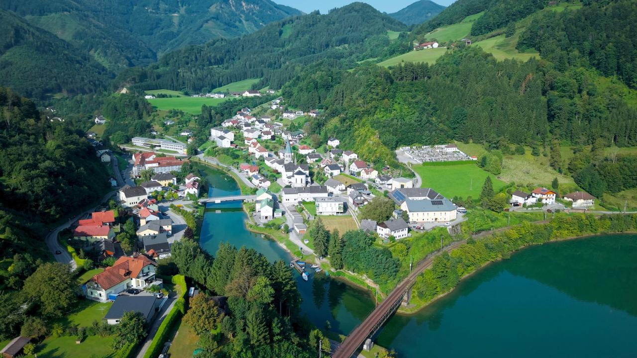 Das Bild zeigt eine malerische Landschaft in der Region Trattenbachtal, insbesondere die Ortschaft Reichraming. Im Vordergrund fließt der Trattenbach, der sich durch die grüne Umgebung schlängelt. Auf der linken Seite des Bildes sind mehrere Häuser mit roten und dunklen Dächern zu sehen, die von Bäumen und Wiesen umgeben sind. 

In der Mitte des Bildes befindet sich eine Brücke, die über den Fluss führt. Auf der rechten Seite sind weitere Gebäude und ein Friedhof erkennbar. Die Landschaft wird von sanften Hügeln und bewaldeten Bergen im Hintergrund umrahmt. Die gesamte Szenerie vermittelt eine ruhige, naturnahe Atmosphäre und zeigt die enge Verbindung zwischen der Siedlung und der umgebenden Natur.