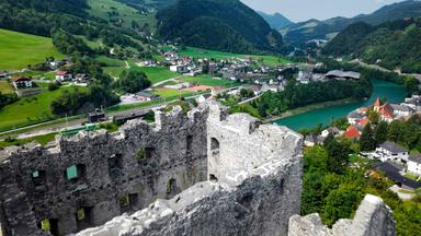 Das Bild zeigt die Ruine der Burg Losenstein, die auf einem Hang in einer malerischen Landschaft liegt. Die Burg hat massive, steinerne Mauern und einige Fensteröffnungen, die eine beeindruckende Aussicht bieten. Im Vordergrund ist der zerfallene Teil der Burg deutlich zu sehen, mit unregelmäßigen Kanten und vertrocknendem Mauerwerk.  Im Hintergrund erstreckt sich eine weite Hügellandschaft, die grün und bewaldet ist. Zwischen den Hügeln verlaufen ein Fluss und mehrere Siedlungen, die aus kleinen Häusern mit roten und weißen Dächern bestehen. Einige Straßen sind sichtbar, darunter eine Eisenbahnlinie, die entlang des Wassers verläuft.   Der Himmel ist teilweise bewölkt, und die Landschaft strahlt Ruhe und Naturverbundenheit aus. Die gesamte Szene vermittelt ein Gefühl von historischer Bedeutung und natürlicher Schönheit.