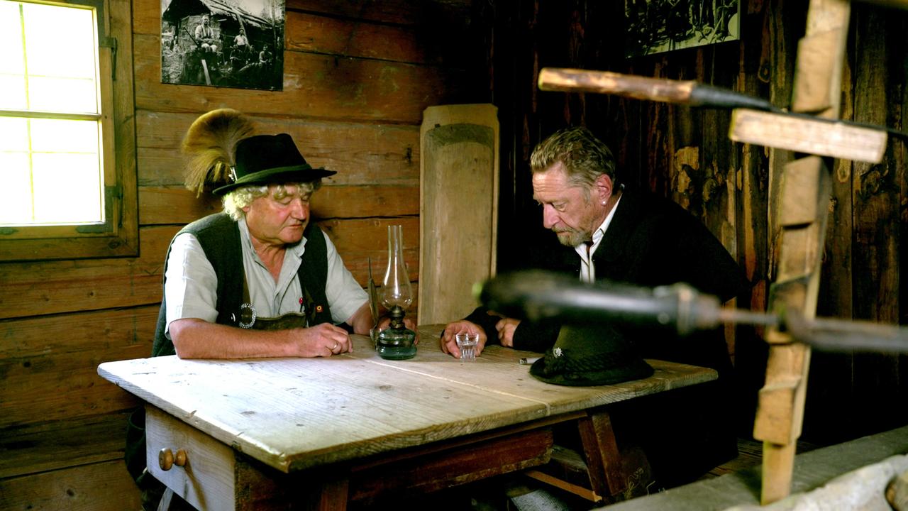 Das Bild zeigt zwei Männer, die an einem rustikalen Holztisch sitzen. Der Tisch ist aus grobem, behandeltem Holz. Neben ihnen steht eine alte Petroleumlampe, die nicht brennt. 

Der Mann links trägt ein traditionelles Outfit, das aus einer hellen Bluse und einer Weste besteht, und hat einen schwarzen Hut mit einem Federbusch auf dem Kopf. Er hat lockige, blond-graue Haare und eine entspannte Körperhaltung.

Der Mann rechts trägt eine dunkle Jacke und hat einen Bart. Er schaut nachdenklich auf den Tisch und hat seine Hände zusammengelegt. 

Im Hintergrund sind Holzwände sichtbar, und an der Wand hängt ein altes Schwarz-Weiß-Foto. Ein Fenster an der linken Seite lässt viel Tageslicht rein und beleuchtet den Raum. 

In der Nähe des Tischs sind einige Werkzeuge sichtbar, die mit Handwerk in Verbindung stehen, was auf eine Umgebung hinweist, die von Tradition und handwerklichen Fertigkeiten geprägt ist.