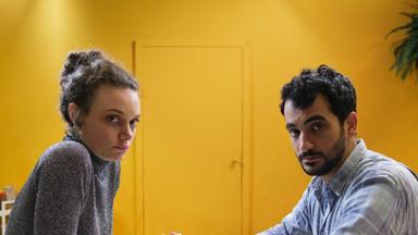 Nelly (Alina Heipe) sitzt mit einem bärtigen jungen Mann, Mervan (Timo Fakhravar) an einem kleinen runden Tisch. Auf dem Tisch liegt ein Küchenmesser. 