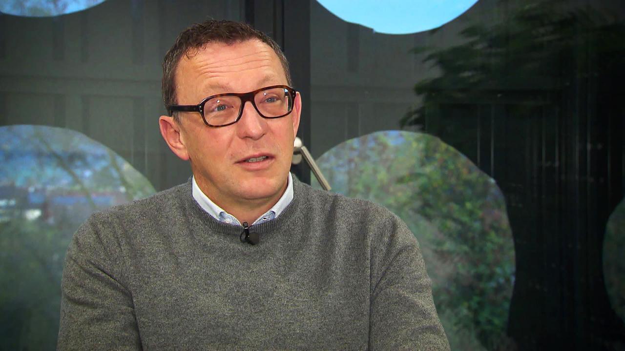 Das Bild zeigt einen Mann mit Brille, der in einem grauen Pullover spricht. Im Hintergrund sind kreisförmige Fenster und Pflanzen sichtbar. Es handelt sich um eine interviewähnliche Situation.