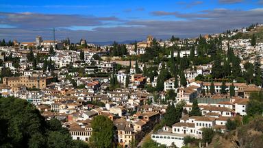 Blick über weiße Häuser mit roten Ziegeldächern an einem grünen Hang in Granada. Zwischen den Gebäuden ragen zahlreiche Zypressen empor.