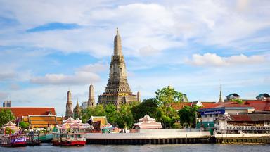 Blick über den Chao Phraya auf den Tempel Wat Arun in Bangkok. Der hohe zentrale Turm ragt über die Tempelanlage und die Gebäude am Flussufer.