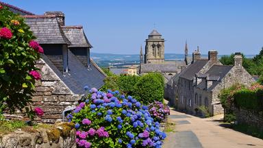 Blick durch eine Straße im mittelalterlichen Dorf Locronan in der Bretagne. Historische Steinhäuser und blühende Hortensien säumen den Weg, dahinter erhebt sich ein Kirchturm.